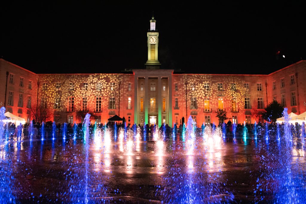 Fellowship Square at night, with the water fountains lit and light projections on the walls of the Town Hall