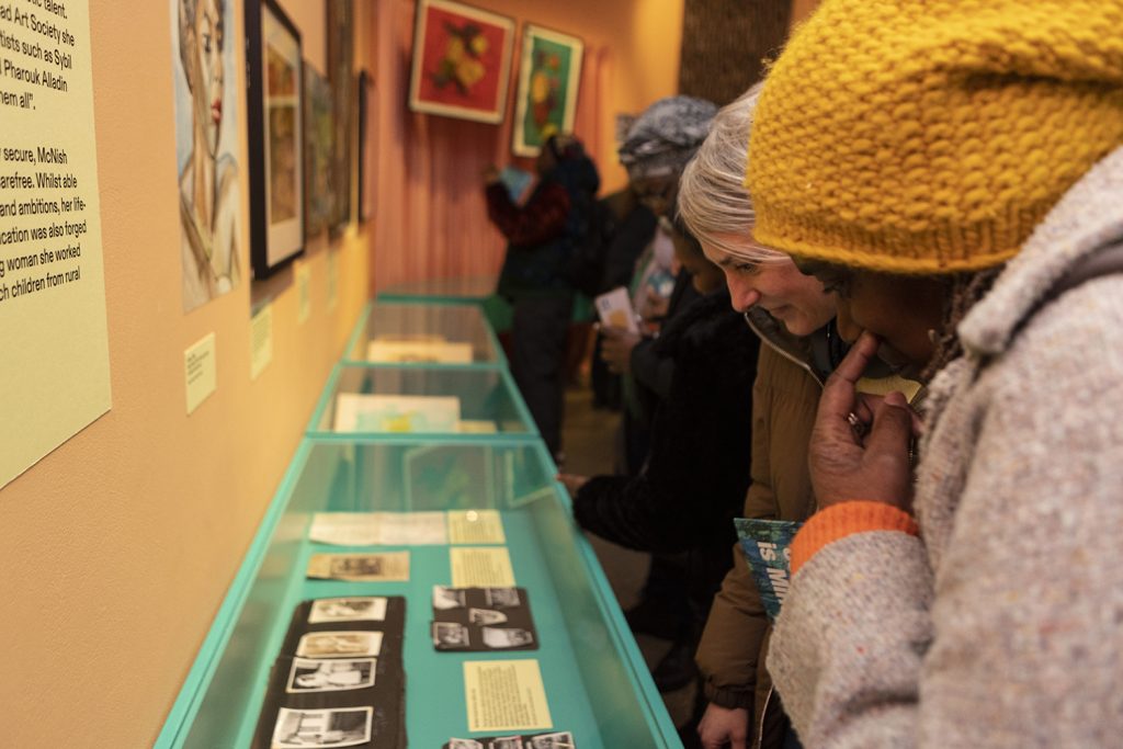 Images of visitors to an exhibition looking down at exhibits in display cases. Photographs can be seen within the cases.
