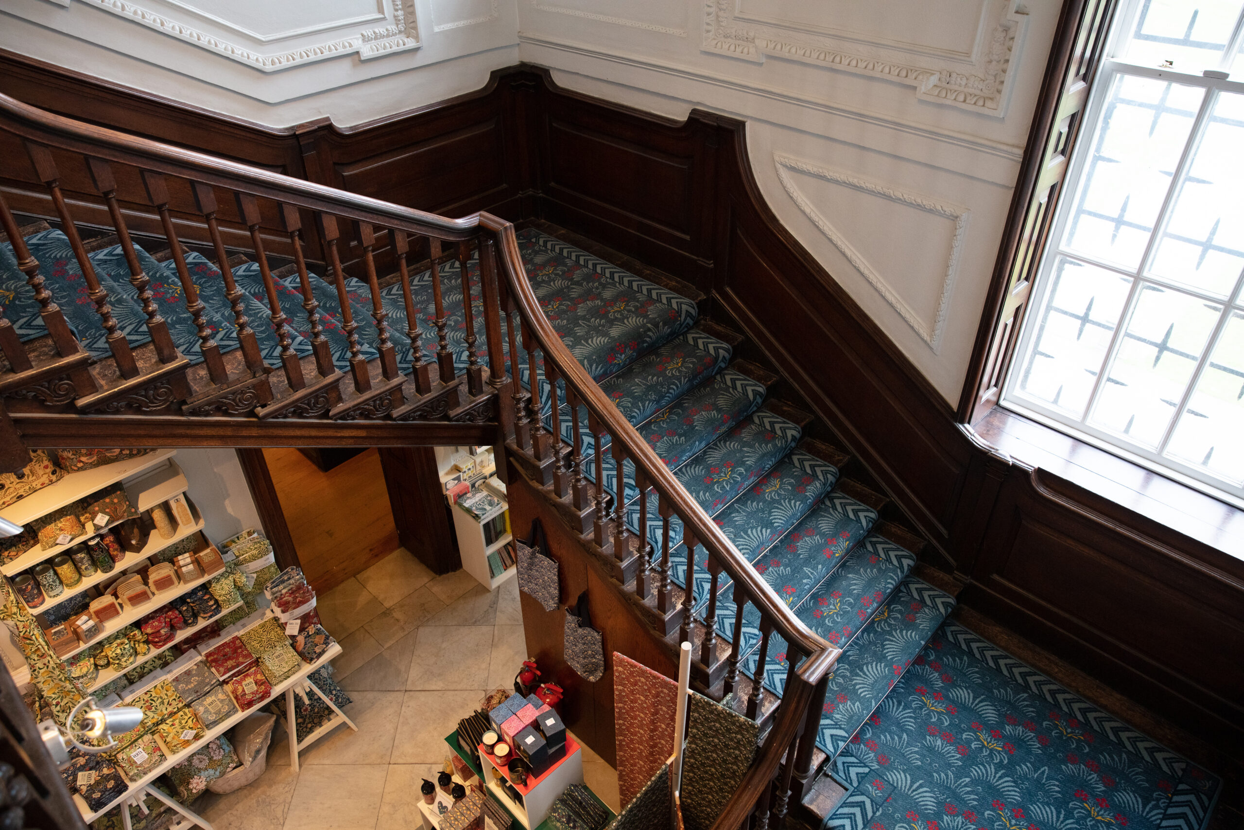 Staircase in William Morris Gallery covered in a blue Morris-design carpet with wooden bannisters, looking down at the shop