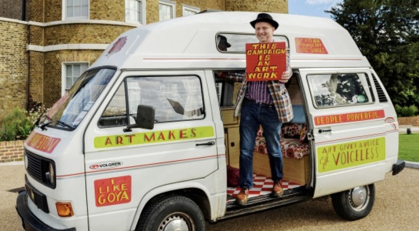 A an in a hat stands in the door of a white van with Art Maker written on the side of it. He holds a banner which reads This campaign is an artwork