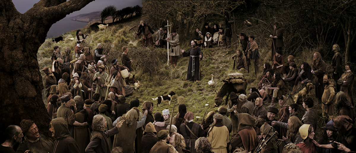 A large crowd dressed in peasant robes gather on a grassy hillside to listen to a speaker